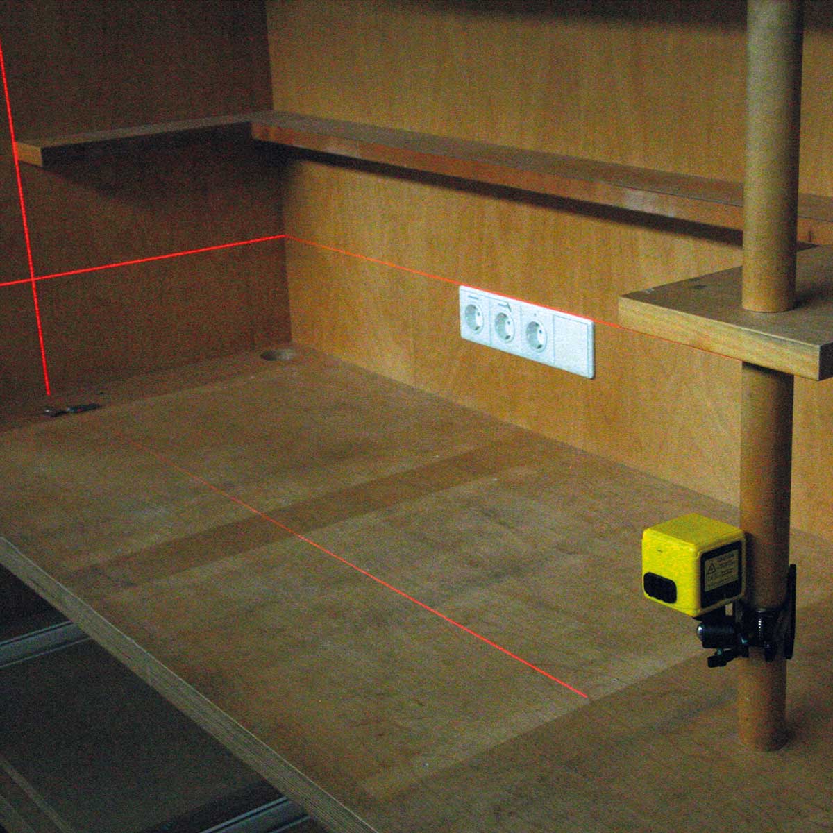 Kreuzlinienlaser an einer Holzstange projiziert rote Linien auf eine Werkbank mit Steckdosen.