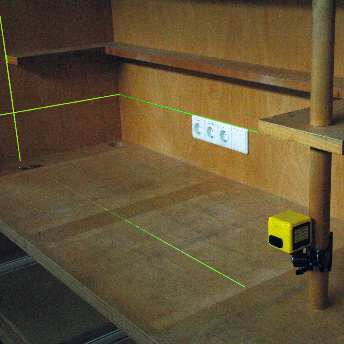 Kreuzlinienlaser an einer Holzstange projiziert grüne Linien auf eine Werkbank mit Steckdosen.