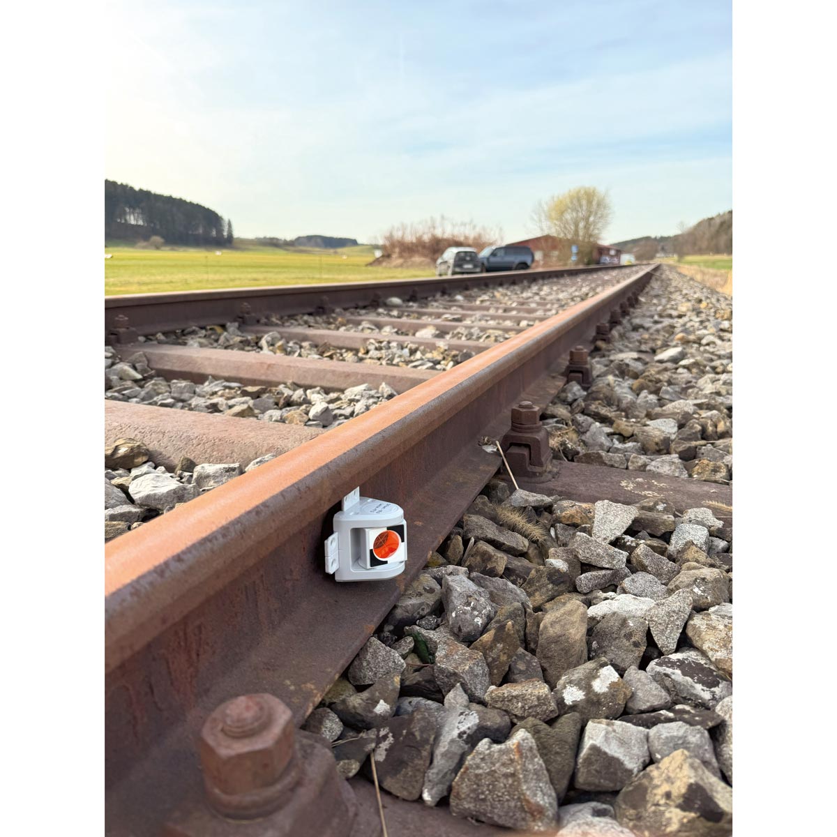 Graues Monitoring-Prisma RSMP380 an einer Bahnschiene im Schotterbett, Blick entlang des Gleises.