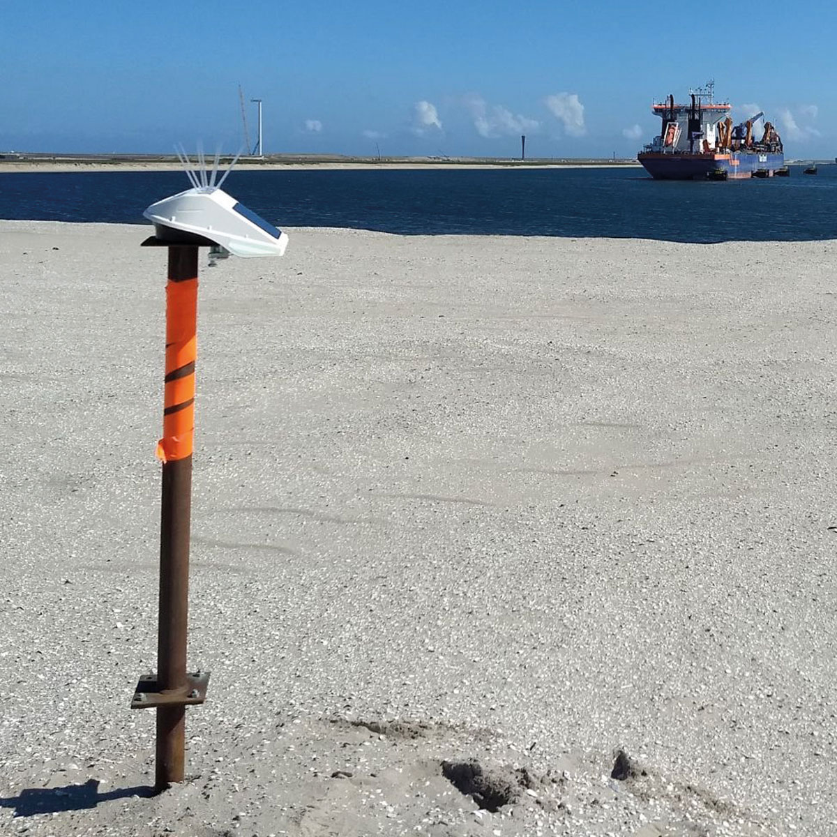 Locator One Sensor auf einer orange-braunen Stange auf einer hellgrauen Sandfläche, mit Schiff im Hintergrund.