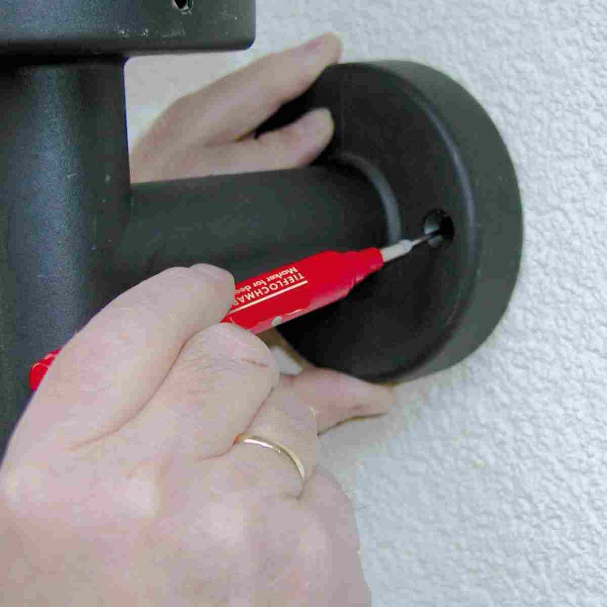 Hand markiert mit dem roten Tieflochmarker ein Bohrloch durch eine Montageplatte an einer Wand.