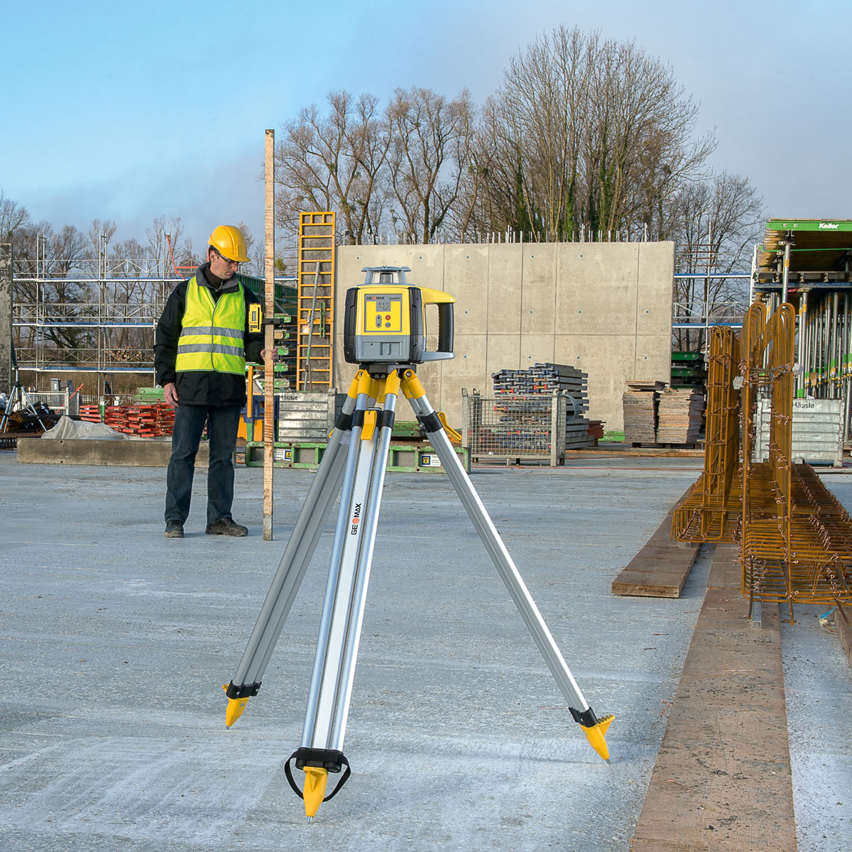 GeoMax Rotationslaser Zone20 H auf Stativ auf einer Baustelle, dahinter Arbeiter mit Messlatte und Empfänger.