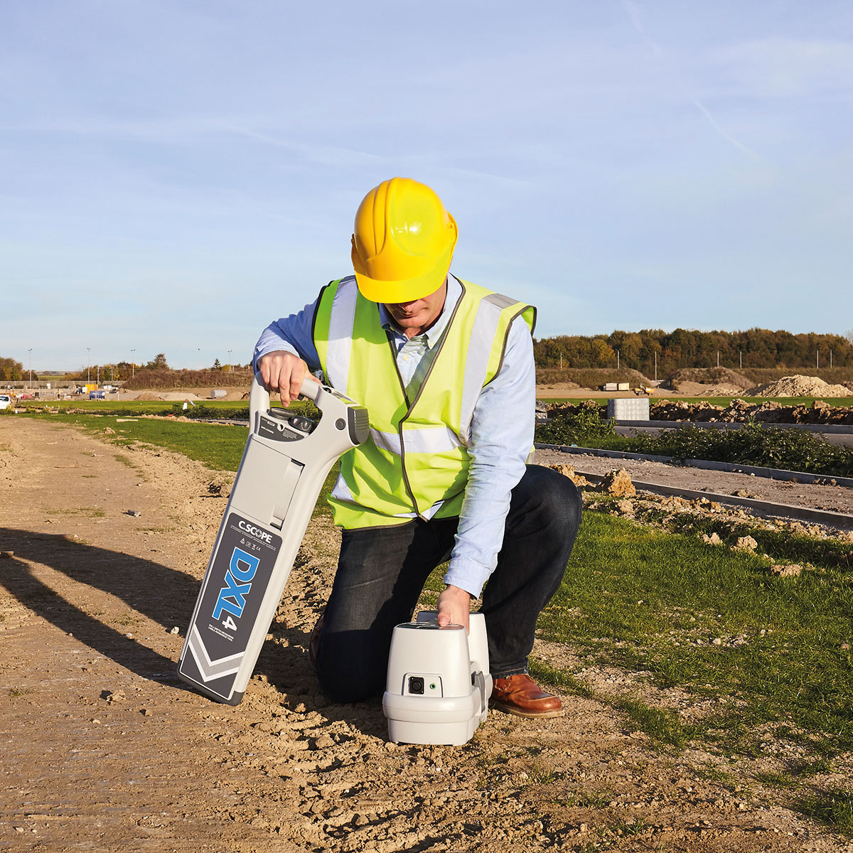 Mitarbeiter mit Helm und Warnweste kniet mit C.Scope Sender SGV4 und Kabelsuchgerät DXL4 auf einer Baustelle.