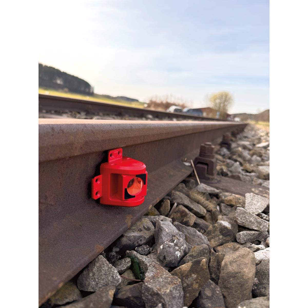 Das rote Monitoring-Prisma RSMP380 von Rothbucher an der Schiene befestigt, mit Schotterbett und Bahngleis.