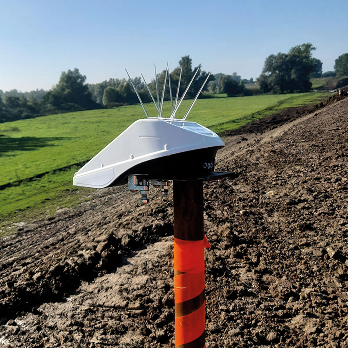 Der Locator One GNSS-Sensor auf einer orange-braunen Stange in einem frisch gepflügten Feld unter blauem Himmel.