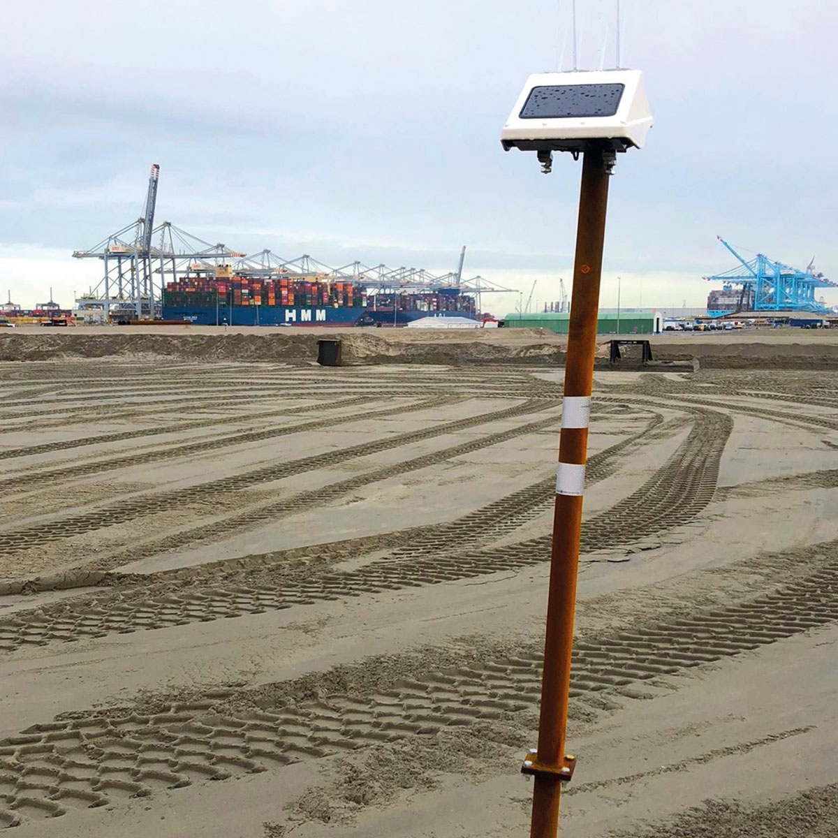 Locator One GNSS auf einer Stange am sandigen Strand mit Reifenspuren und maritimen Strukturen im Hintergrund.